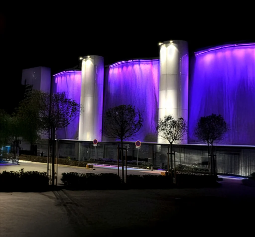 Façade architecturale éclairée en violet la nuit avec colonnes lumineuses et arbres au premier plan